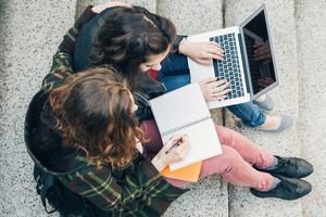 Two university students taking notes on a paper notebook and a laptop