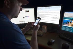 A screenshot of a man sitting in front of three computers and using his phone